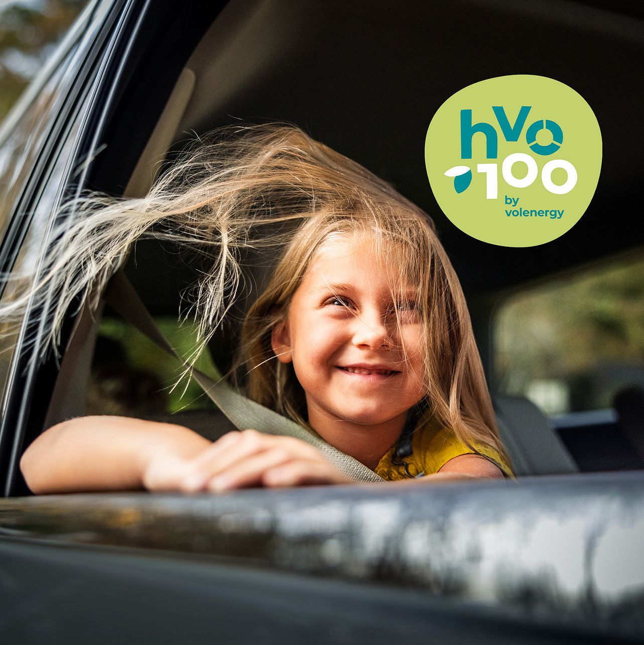 Fröhliches Kind mit flatterndem Haar lehnt sich aus dem geöffneten Autofenster, angeschnallt auf dem Rücksitz – daneben das grüne Logo von HVO100 by Volenergy als Zeichen für nachhaltige Mobilität.