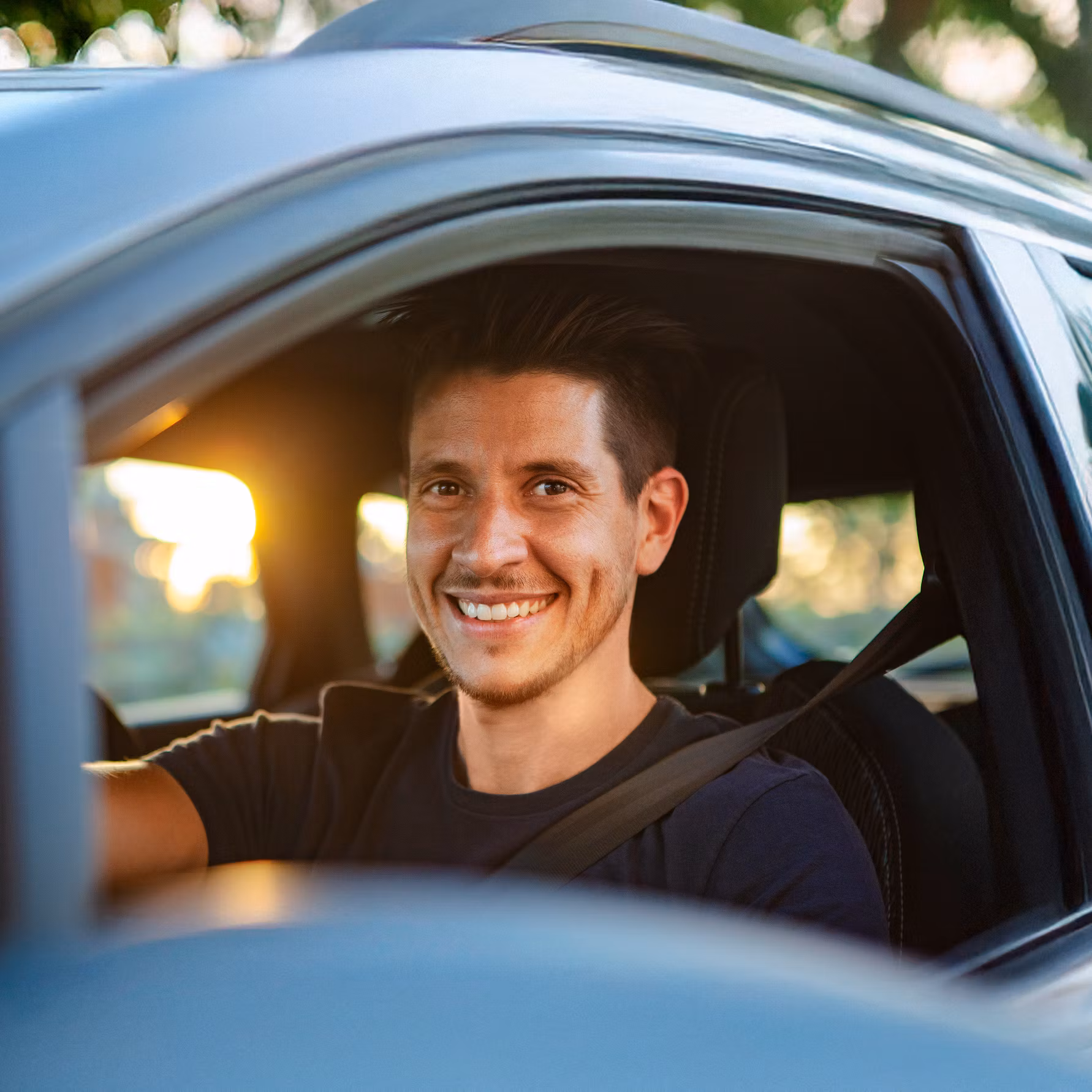 Lächelnder Mann am Steuer eines Autos, mit geöffnetem Fenster und angeschnallt, im Hintergrund Bäume in warmem Sonnenlicht – symbolisiert Freude am Fahren und nachhaltige Mobilität