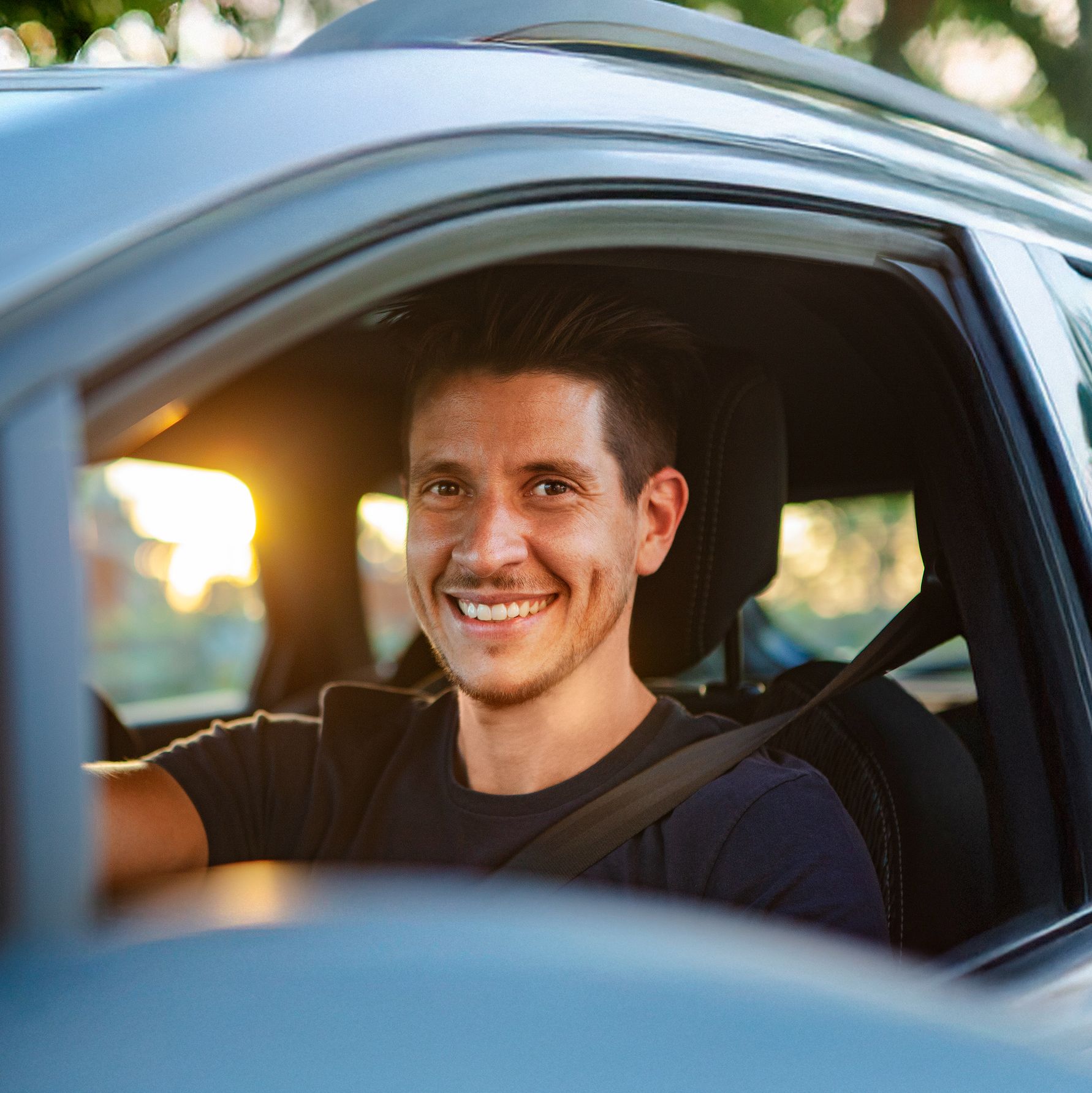 Lächelnder Mann am Steuer eines Autos, mit geöffnetem Fenster und angeschnallt, im Hintergrund Bäume in warmem Sonnenlicht – symbolisiert Freude am Fahren und nachhaltige Mobilität