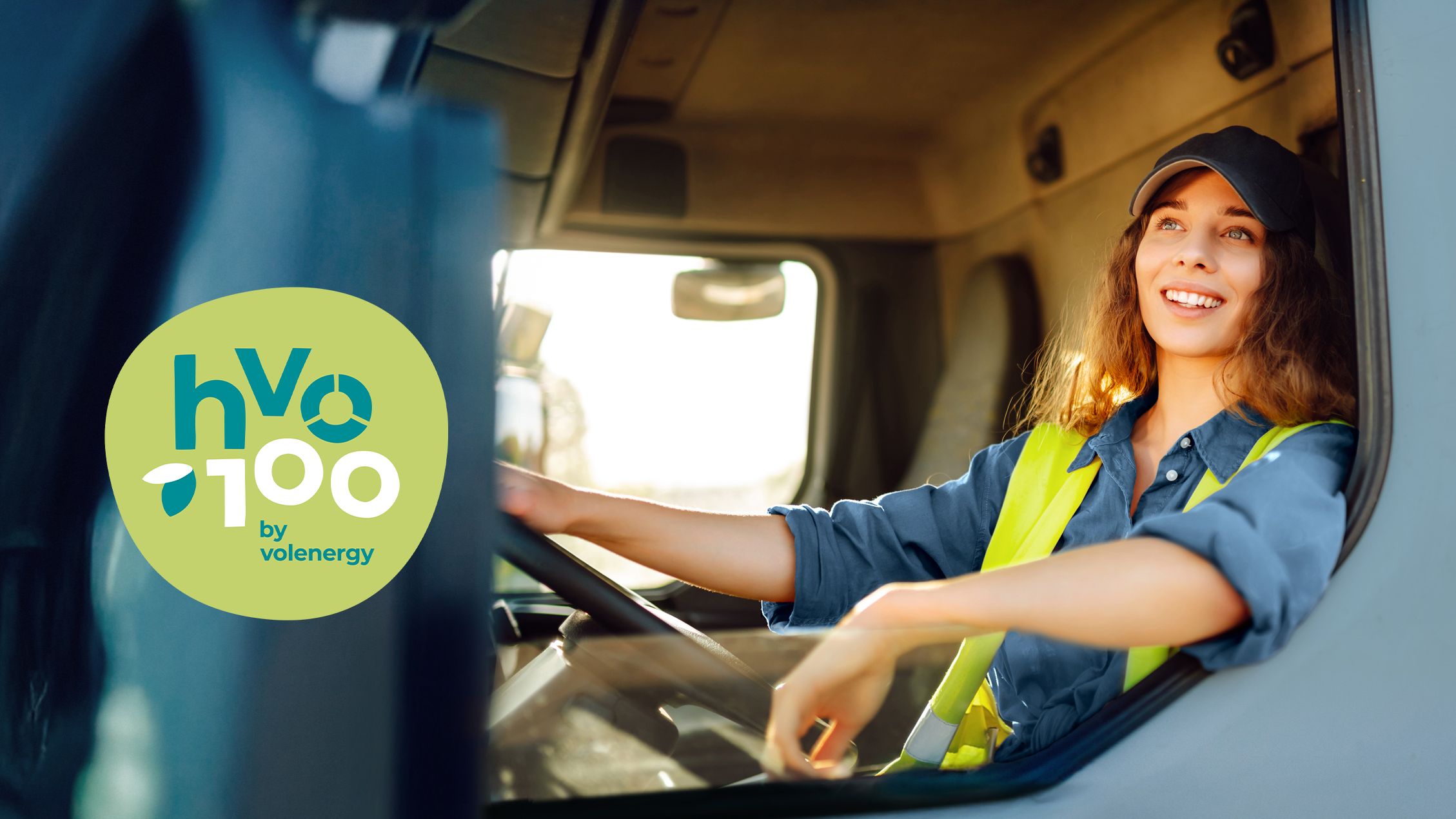 Junge Frau mit Sicherheitsweste sitzt lächelnd in der Fahrerkabine eines Lastwagens, links im Bild das grüne Logo von HVO100 by Volenergy – Hinweis auf klimafreundlichen Kraftstoff.