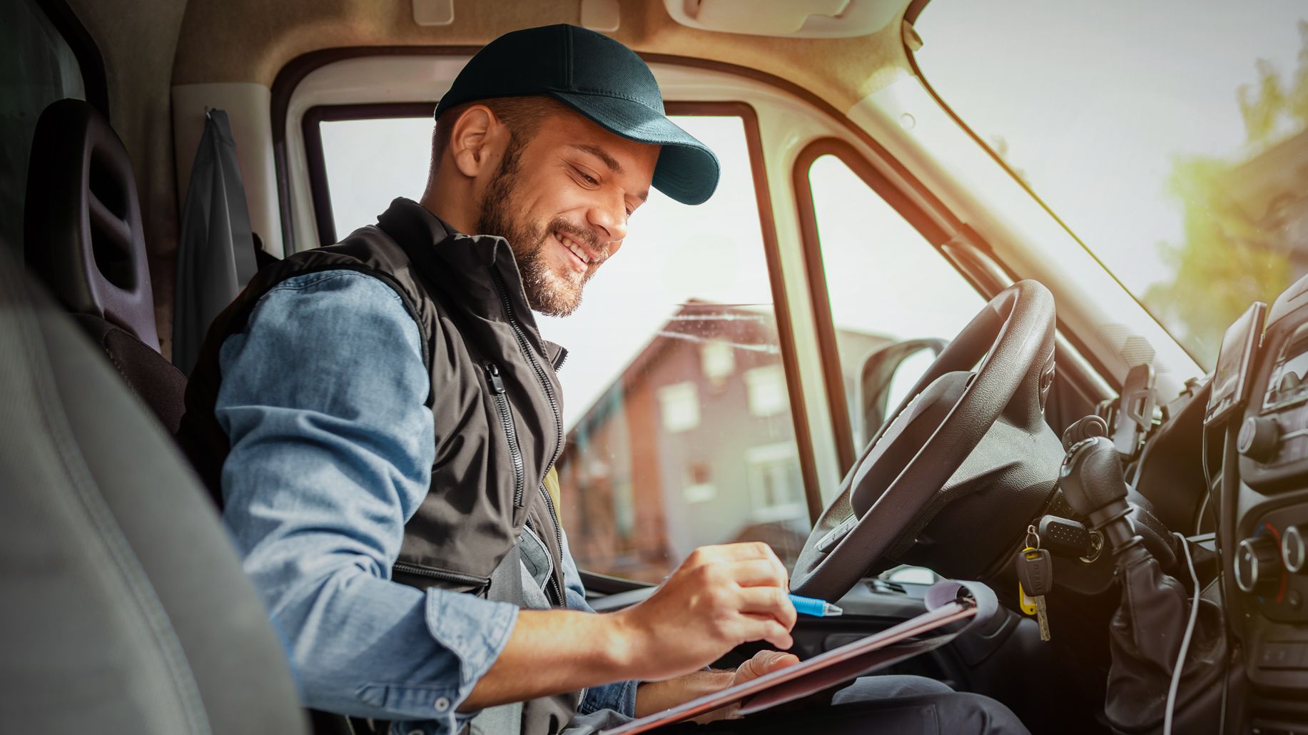 Lieferfahrer mit Basecap sitzt lächelnd im geparkten Transporter und schreibt auf ein Klemmbrett – symbolisiert effiziente und verantwortungsvolle Mobilität im Arbeitsalltag.